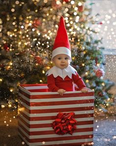 a baby dressed as an elf sitting in a gift box next to a christmas tree