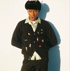 a man in a black sweater and hat standing against a white wall