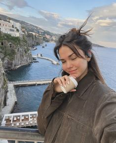 a woman standing on top of a balcony next to the ocean holding a cup in her hand