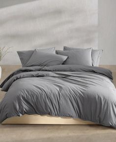 a bed with grey sheets and pillows on top of it in a white room next to a potted plant