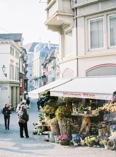 people are walking down the street in front of some buildings with flowers and plants on display