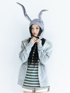 a young woman wearing a bunny ears hat and grey coat standing in front of a white wall