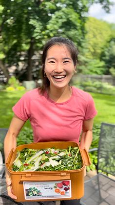 a woman is holding a box full of salads and smiling at the camera while sitting on a patio table