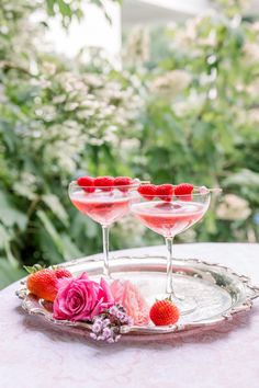 two martinis with strawberries sit on a silver tray in front of some flowers