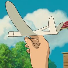 a hand holding a paper airplane in front of a tree and sky with clouds behind it