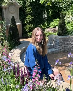 a woman sitting on a bench in the middle of flowers and plants with a clock behind her