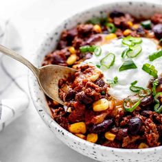 a white bowl filled with chili and corn topped with sour cream on top, next to a spoon