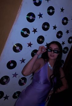 a woman in a purple dress and sunglasses posing for the camera with cds on the wall behind her