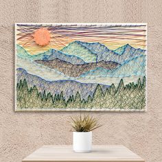 a wall hanging above a table with a potted plant on it and mountains in the background