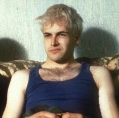 a man with blonde hair sitting on a couch in front of a blue wall and wearing a blue tank top