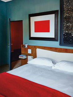 a bed with white sheets and red blanket in a blue room next to a painting on the wall