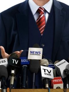 a man in a suit and tie standing behind microphones with news channel logos on them