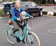 a woman riding a bike with her dog on the back in front of them and cars behind her