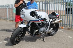 two men standing next to a white and blue motorcycle in parking lot with fence behind it