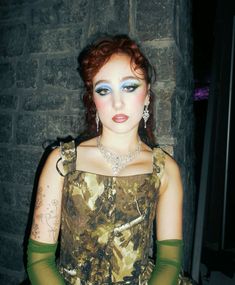 a woman with red hair and blue eyes wearing green gloves is posing in front of a brick wall