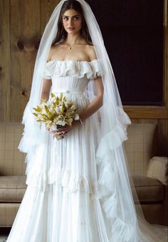 a woman in a white wedding dress standing next to a couch and holding a bouquet