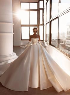 a woman in a white wedding dress standing by a window with her hands on her hips