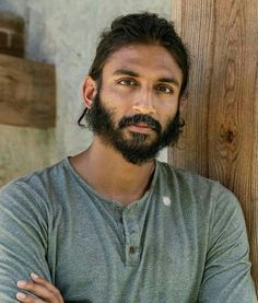 a man with long hair and beard standing in front of a wooden wall wearing a gray shirt