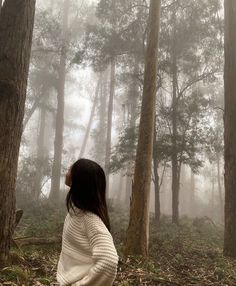 a woman standing in the middle of a forest on a foggy day with trees