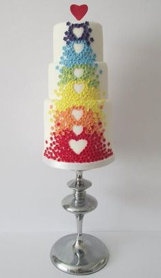 a multi - colored cake with hearts on top is sitting on a silver stand and has a white base