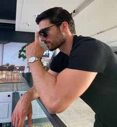 a man in sunglasses leaning on a railing looking at his watch while holding his hand to his face