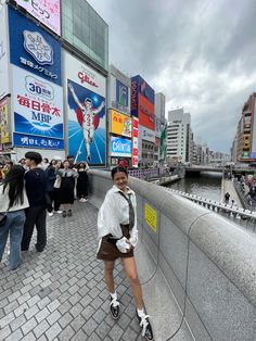 a woman standing on the side of a river next to tall buildings and billboards