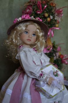 a doll with blonde hair wearing a white dress and pink hat holding a gift box