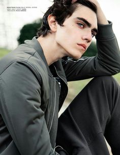 a young man sitting on the ground with his hand in his hair and wearing a jacket