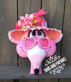 a pink elephant wearing sunglasses and a hat with flowers on it's head, standing next to a wooden fence