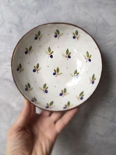 a hand holding a white bowl with blue berries on the bottom and green leaves on it