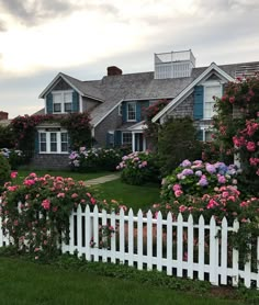 a white picket fence surrounded by pink and purple flowers in front of a gray house