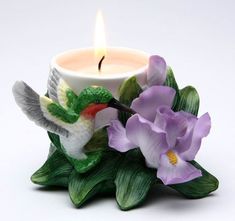 a candle that is sitting on top of some leaves and flowers with a humming bird