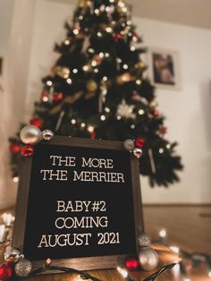 a sign that says the more the merrier baby 2 coming august 2011 in front of a christmas tree