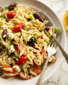 a white plate topped with pasta salad next to a fork and lemon wedges on a marble table