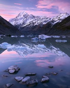 the mountains are covered in snow and ice as the sun sets over water with rocks on it