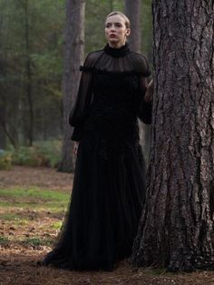 a woman in a black dress standing next to a tree with her hands on her hips