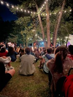 a group of people sitting on the ground in front of a tree with lights strung over it