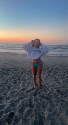 a woman standing on top of a sandy beach next to the ocean with her hands behind her head