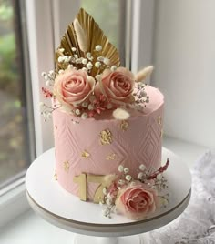 a pink cake decorated with gold leaf and flowers sitting on a table next to a window