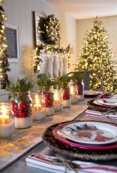 a table set for christmas dinner with lit candles and decorated trees in the back ground