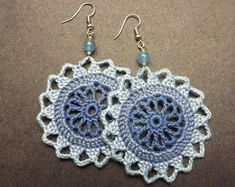 two crocheted doily earrings are shown on a brown surface, one is blue and the other is white