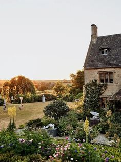 an old house surrounded by lots of flowers and greenery in the sun set on a sunny day