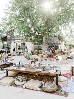 an outdoor dining area with tables, chairs and pillows on the ground in front of a large tree