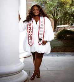 a woman in white graduation gown standing next to a column and smiling at the camera