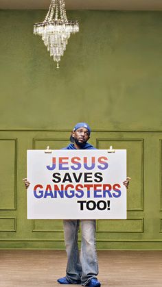 a man holding a sign that says jesus saves gangsters too in front of a green wall