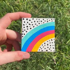 a hand holding up a small piece of paper with a rainbow painted on it in the grass