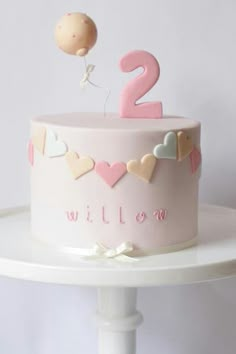 a pink two year old birthday cake with hearts and balloons on the top, sitting on a white pedestal