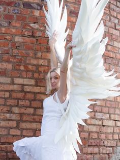 a woman in white dress holding up large white wings next to brick wall with bricks behind her