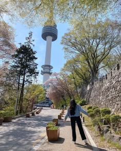 a woman standing on the side of a road next to a tall tower with a sky scraper in the background