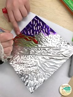 a person is working on an art project with silver foil and colored pencils in front of them